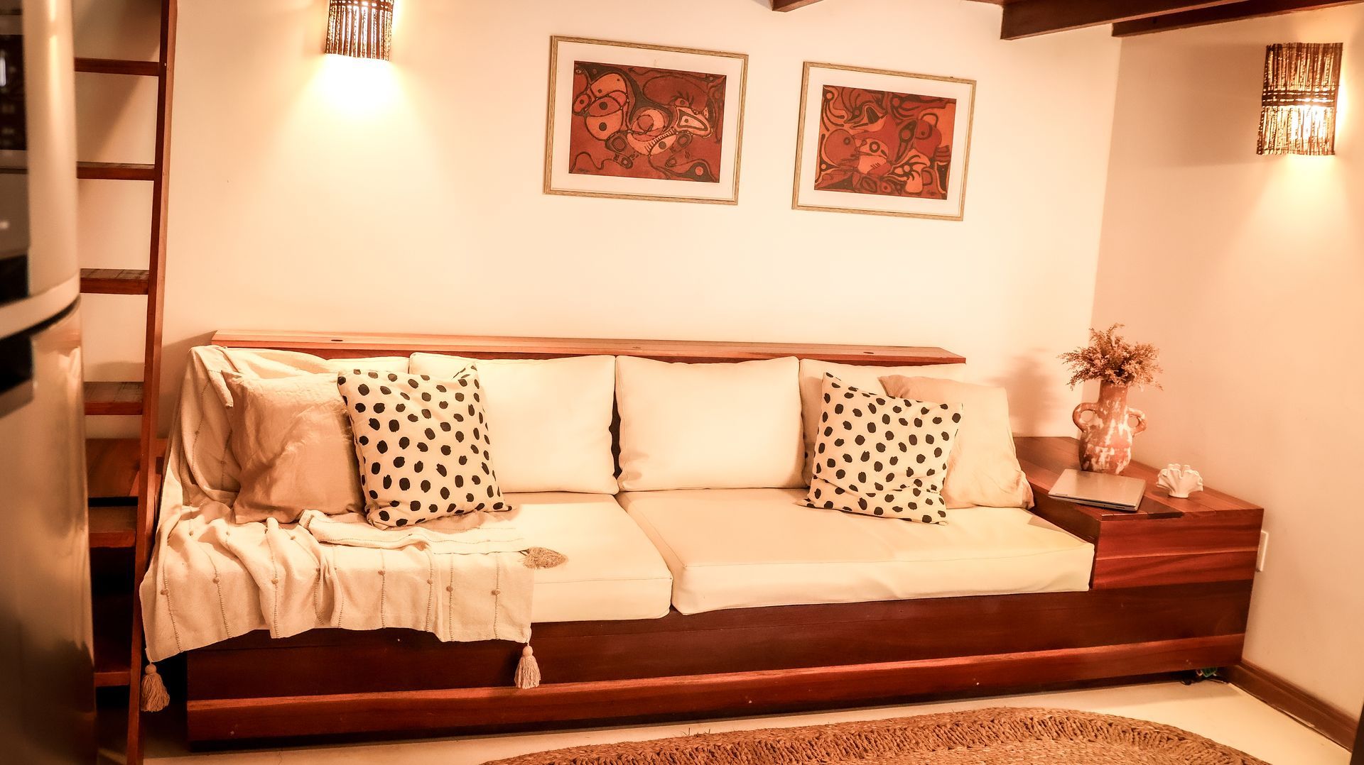 Sofá de madeira com almofadas estampadas contra uma parede branca, obras de arte emolduradas e iluminação aconchegante.