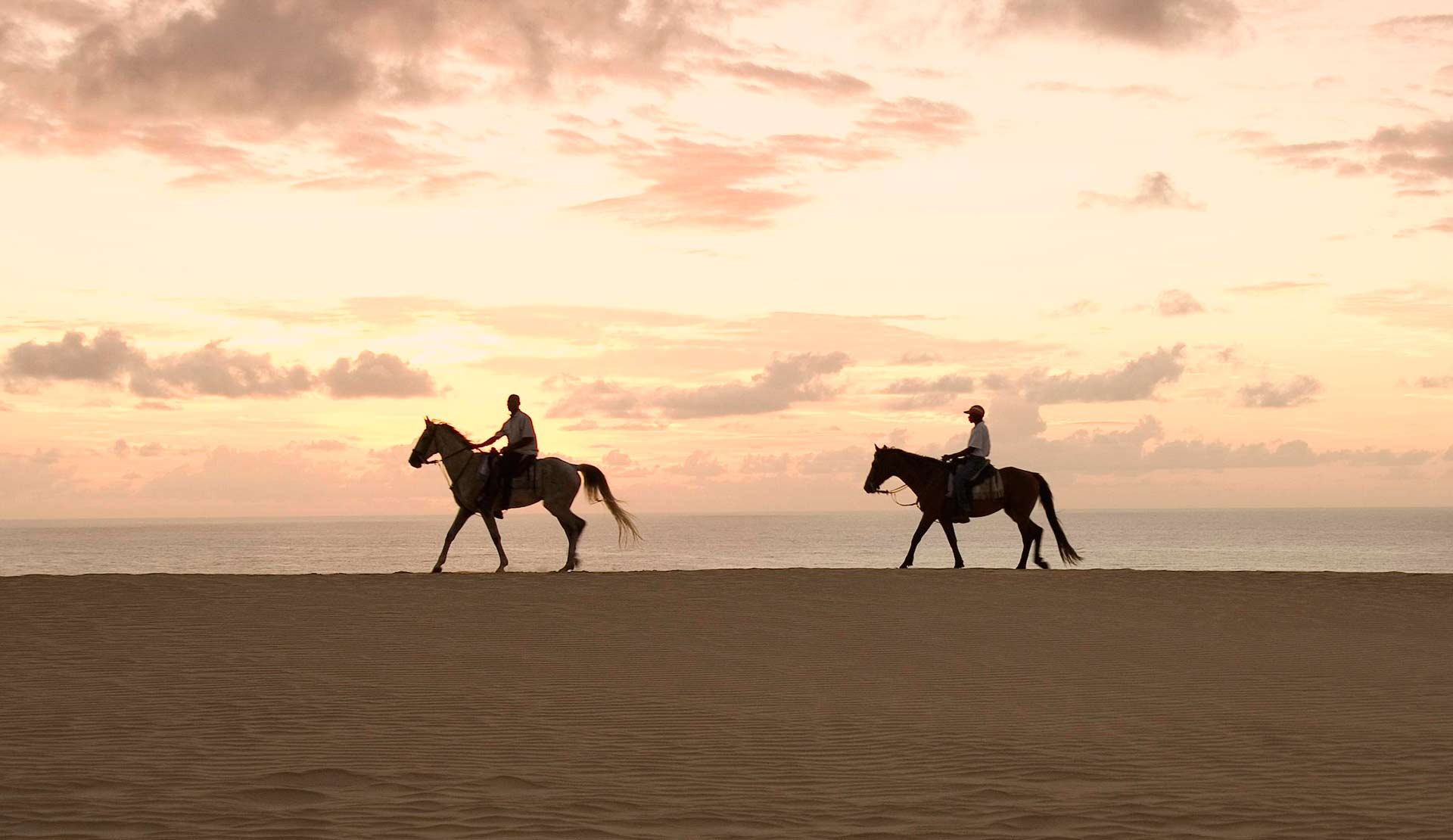 Duas pessoas estão andando a cavalo na praia ao pôr do sol.