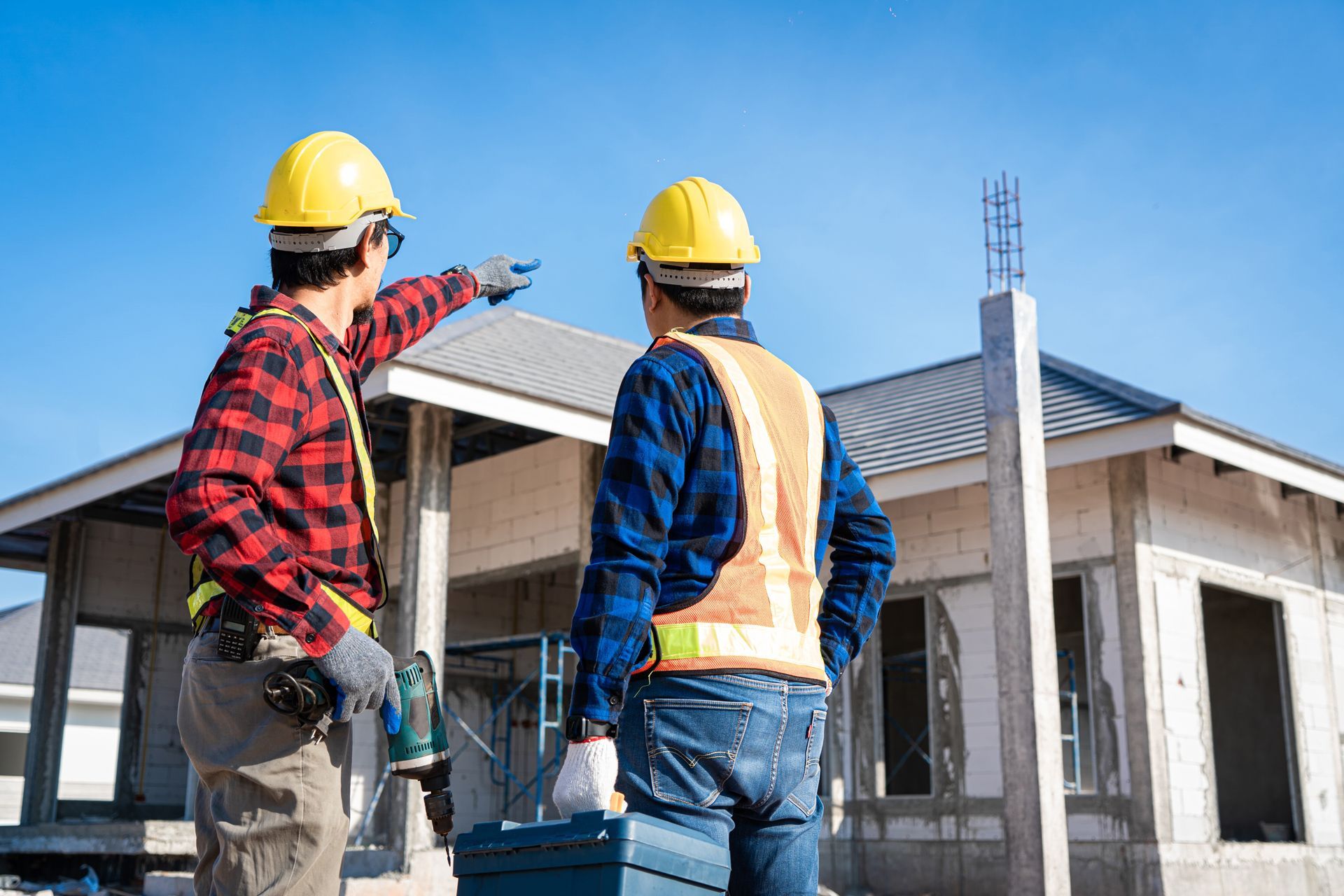 Two construction workers wearing hard hats and vests at a construction site, one pointing, the other holding a toolbox.