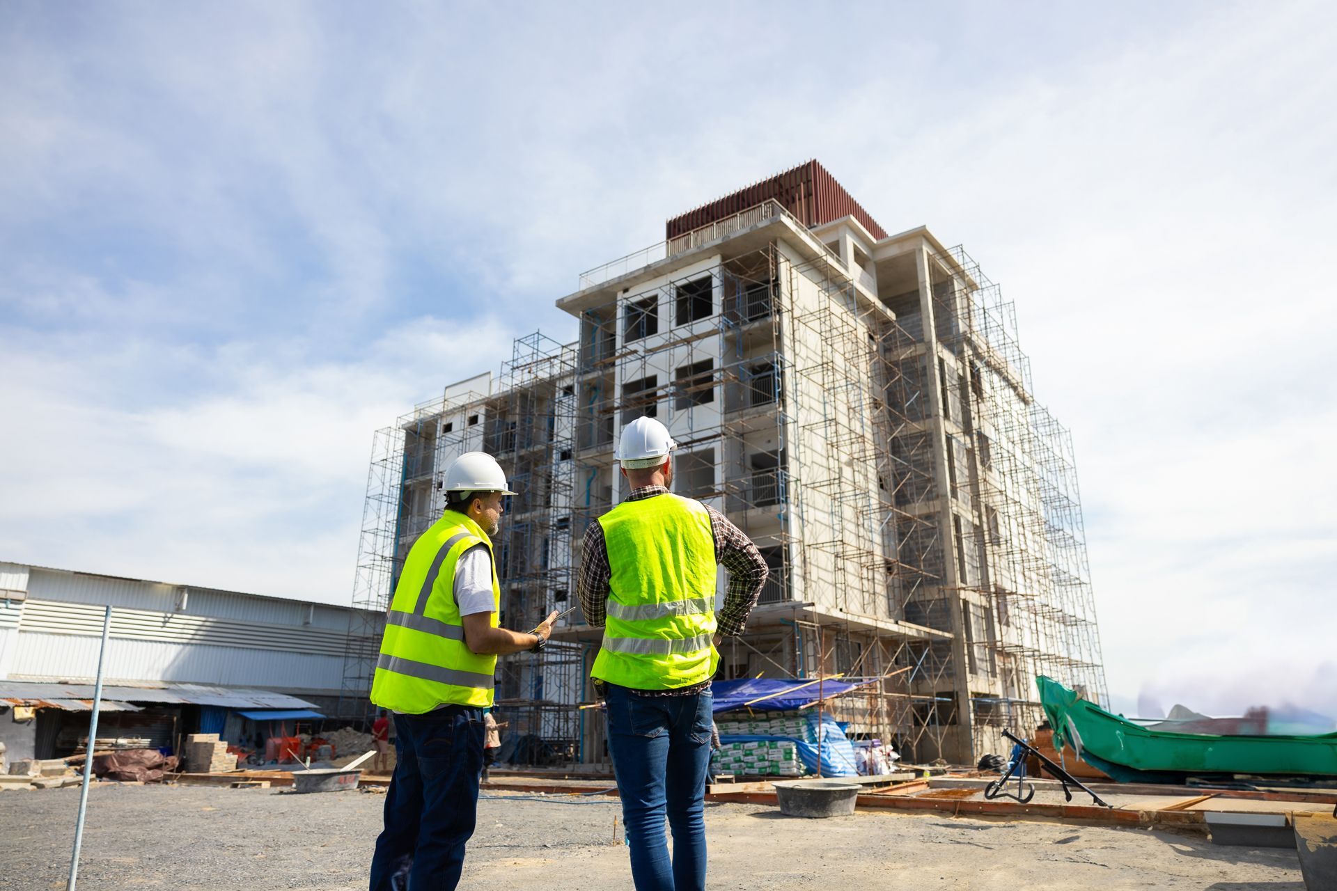 Two construction workers in vests and hard hats discuss plans in front of a building under construction.