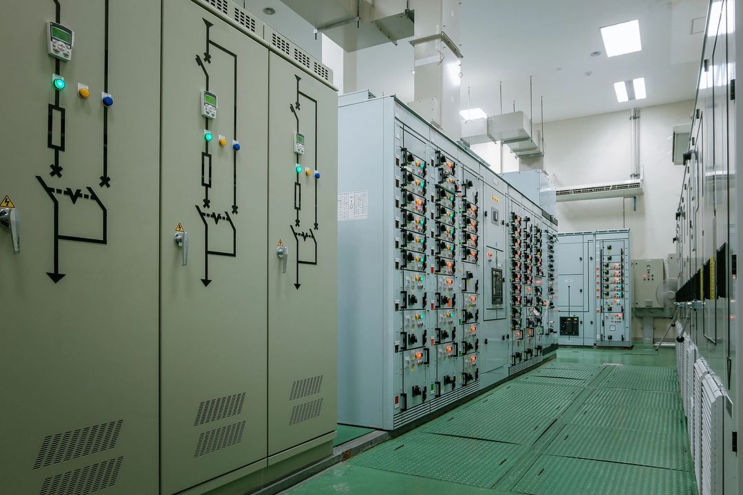 File di grandi armadi elettrici grigi con schemi elettrici in una sala apparecchiature industriale ben illuminata.