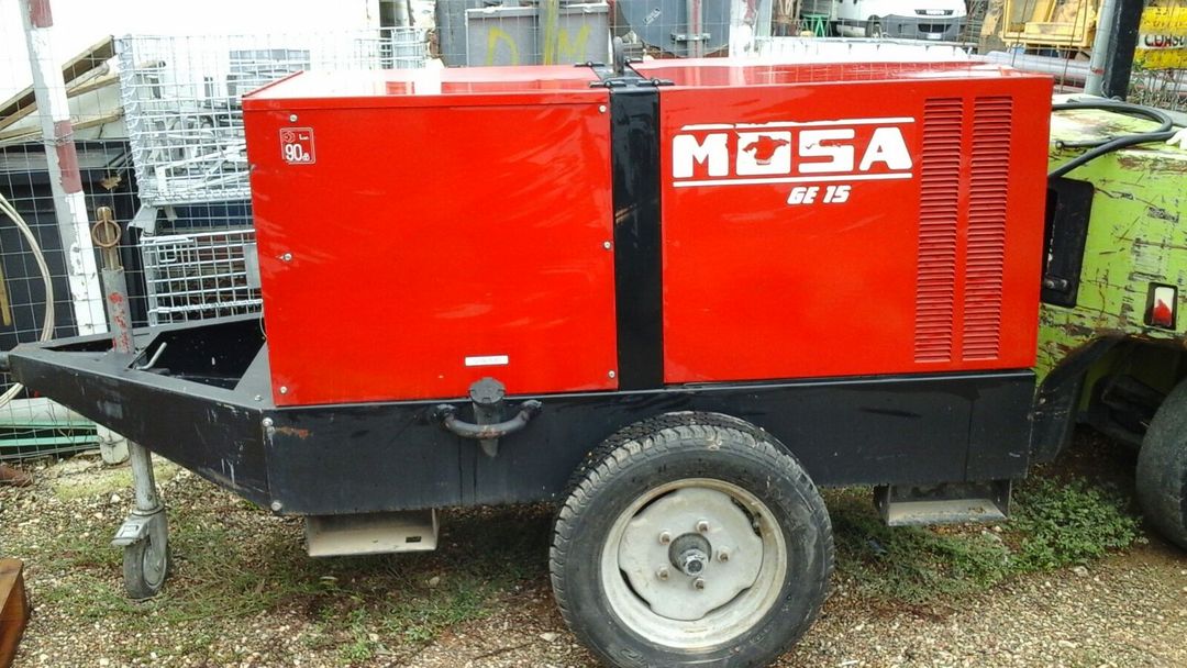Un generatore Mosa GE 15 rosso, montato su un rimorchio metallico nero con una sola ruota, è posizionato in un'area di stoccaggio esterna.