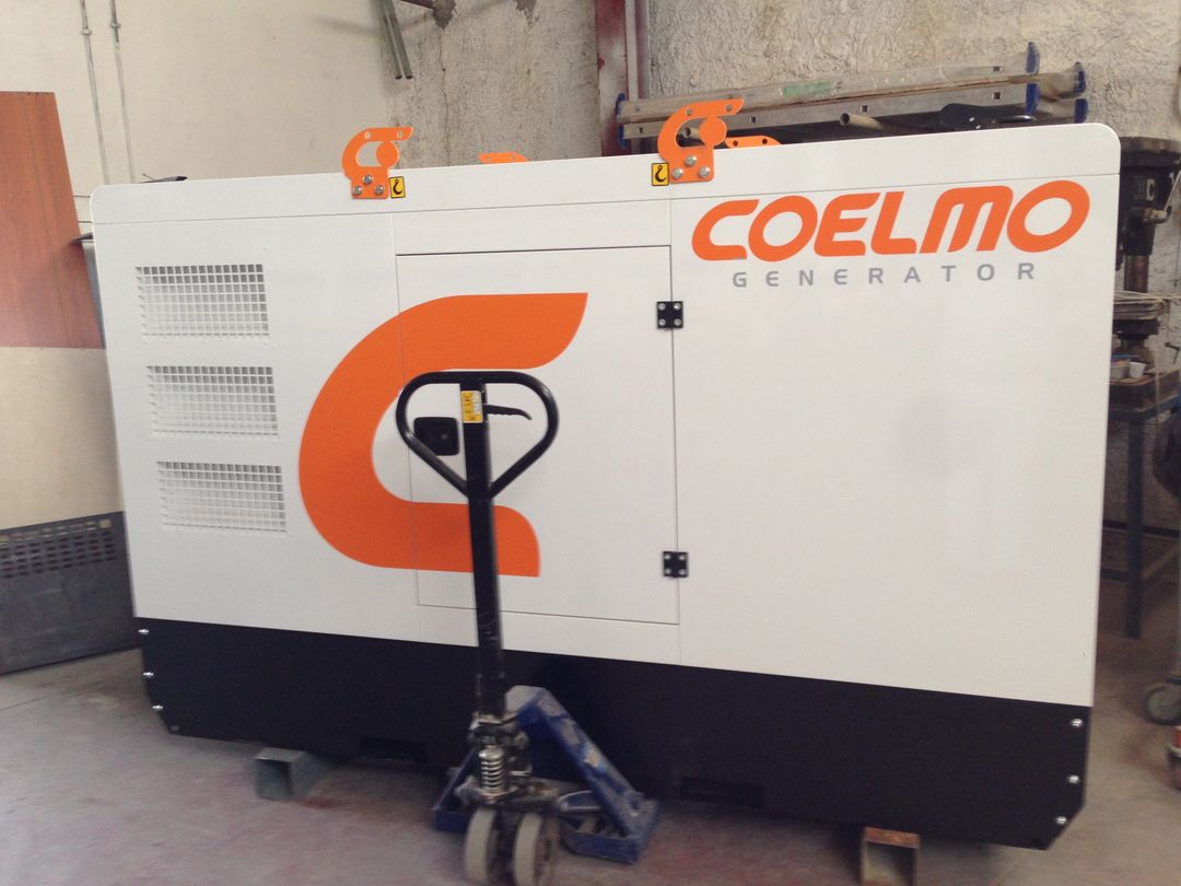 Un generatore di corrente bianco e nero della marca Coelmo con logo arancione, posizionato in un locale con un transpallet manuale davanti.