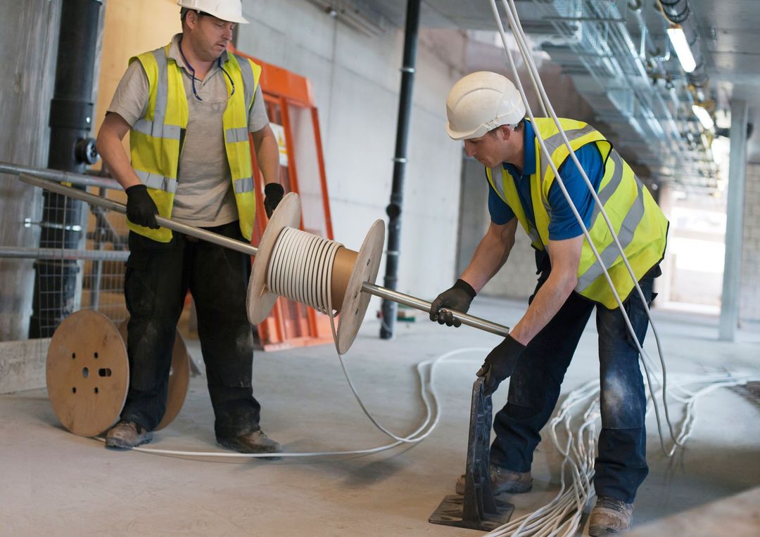 Due operai edili, con indosso giubbotti ad alta visibilità e caschi di sicurezza, lavorano insieme per srotolare un cavo elettrico per impieghi gravosi.