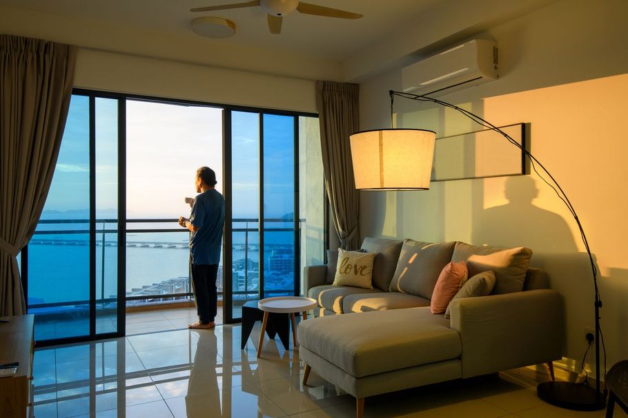 Una persona si trova su un balcone affacciato sull'oceano al tramonto, all'interno di un soggiorno con un divano e una lampada da terra ad arco.