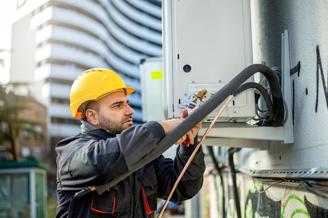 Un tecnico con elmetto giallo e giacca da lavoro installa o effettua la manutenzione di un'unità esterna di condizionamento dell'aria su una parete.