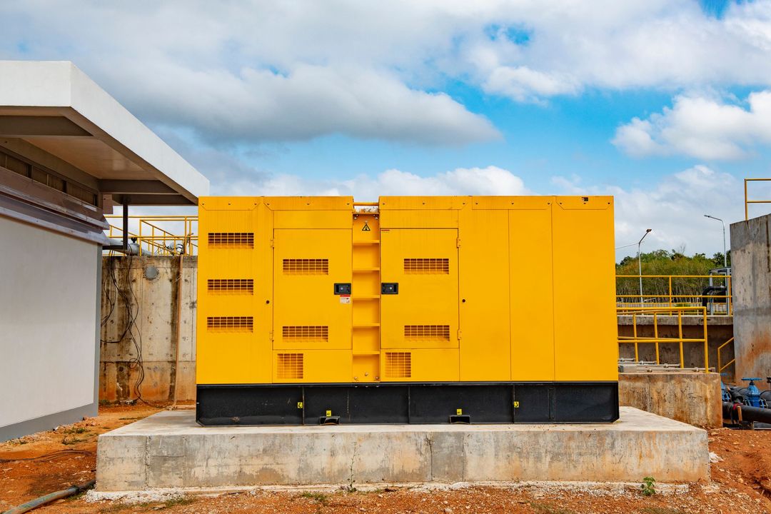 Un grande generatore industriale giallo è posizionato su una base di cemento all'aperto, sotto un cielo azzurro con nuvole.