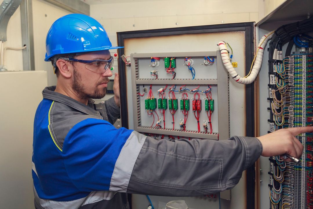 Una persona con un elmetto blu e una divisa da lavoro indica dei fili in un quadro elettrico industriale a vista.