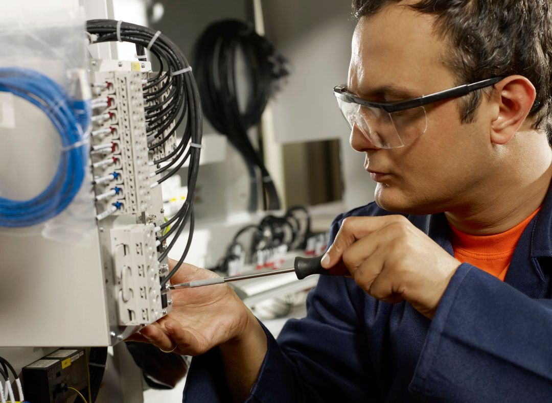 In un laboratorio, una persona con occhiali protettivi utilizza un cacciavite per lavorare su un pannello elettrico bianco con fili neri.