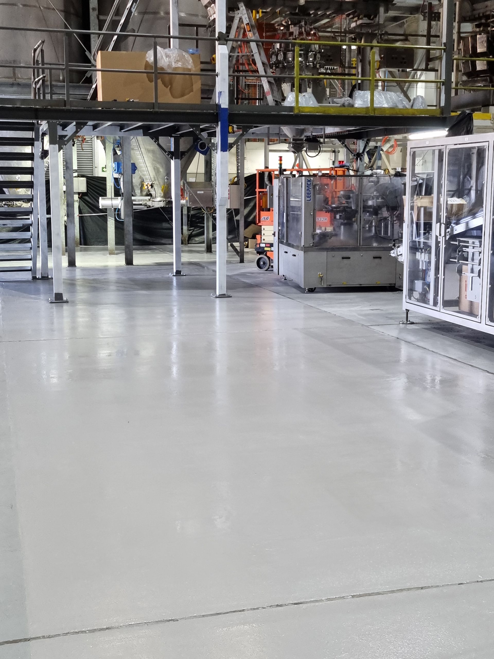 Industrial factory floor with machines and shelving under overhead piping, viewed across a polished concrete aisle
