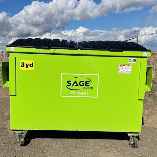 A green dumpster with a black lid is sitting on top of a dirt field.