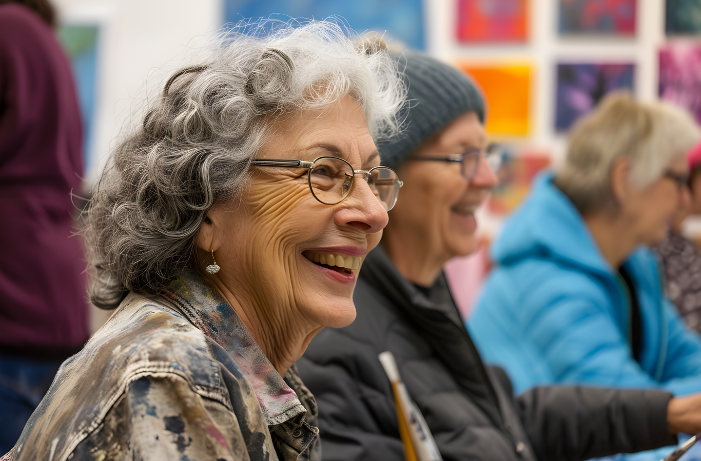 Mujer mayor sonriente con gafas en una clase de arte, rodeada de otras personas, con obras de arte coloridas en el fondo.