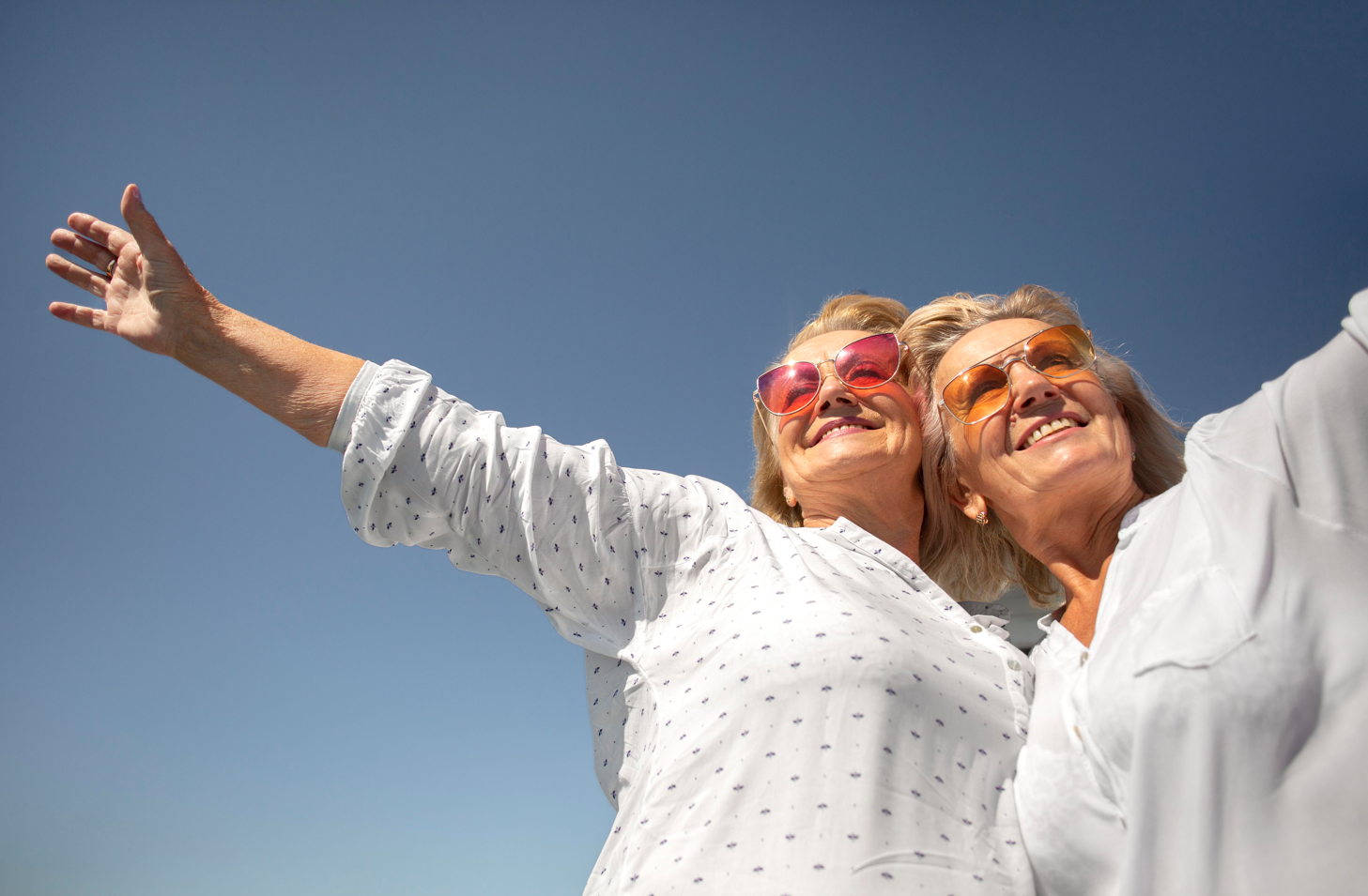 Dos mujeres mayores con camisas blancas y gafas de colores levantan los brazos contra un cielo azul, sonriendo.