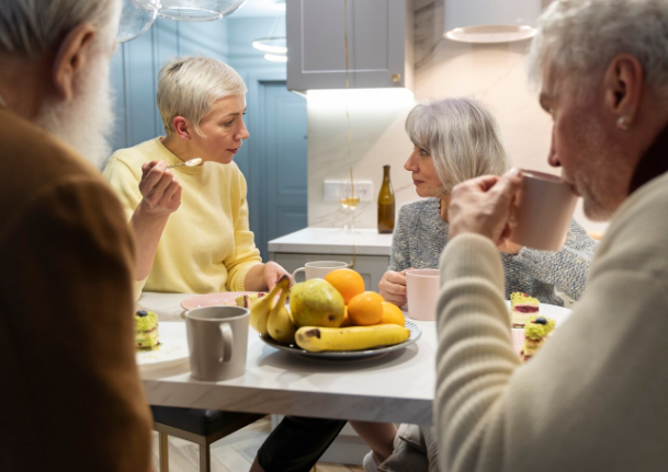 Cuatro adultos mayores en una mesa de cocina con frutas, bebidas y pasteles, conversando.