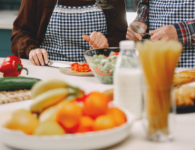 Dos personas cocinan juntas en una cocina, picando verduras. Hay frutas, pasta y una botella de leche sobre la encimera.