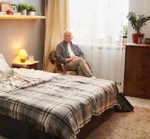 Un hombre mayor se sienta en una silla junto a una ventana de su dormitorio, mirando hacia la cámara.