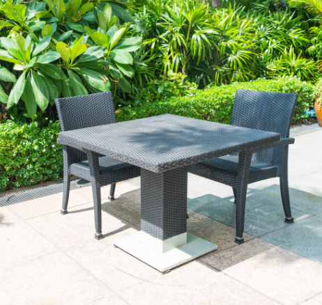 Mesa cuadrada con dos sillas en un patio, rodeada de vegetación. Muebles de mimbre negro, día soleado.
