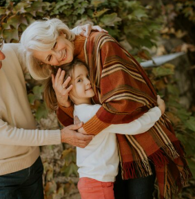 Abuela abrazando a una niña, con su abuelo cerca; colores de otoño, expresiones felices.