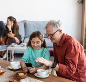 Abuelo y nieta mirando un teléfono juntos en una mesa, otros miembros de la familia en el fondo.