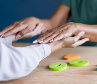Dos manos flotando sobre las manos de otra persona, juguetes verdes y naranjas sobre una mesa de madera.