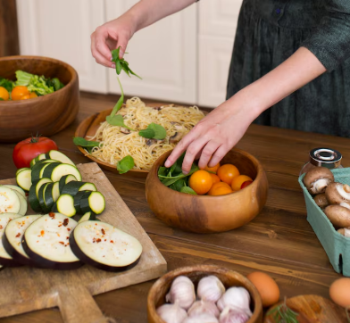 Persona preparando ingredientes en cuencos de madera: verduras, pasta, champiñones y huevos en una mesa de madera.