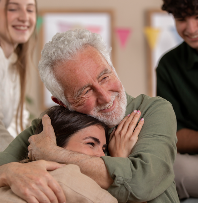 Un hombre mayor sonriente abraza a una joven. Otros sonríen al fondo. Dentro, luz natural.