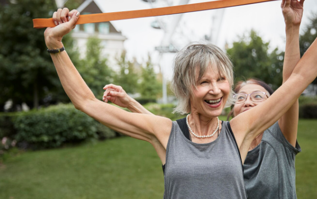 Dos mujeres mayores sonriendo, estirándose con una larga banda naranja al aire libre.