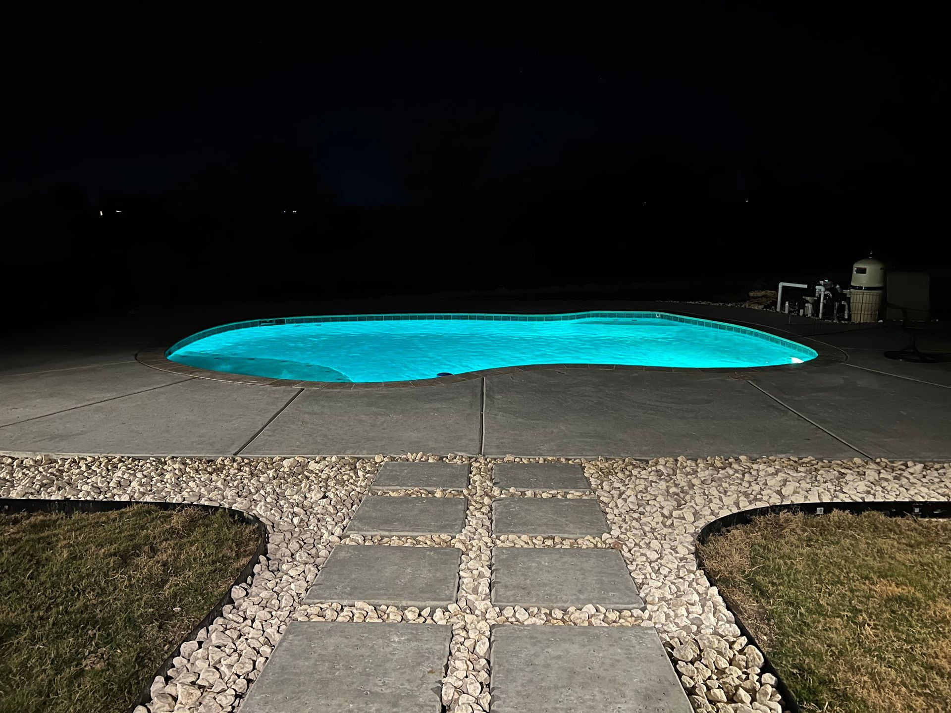 A swimming pool is lit up at night with a walkway leading to it.
