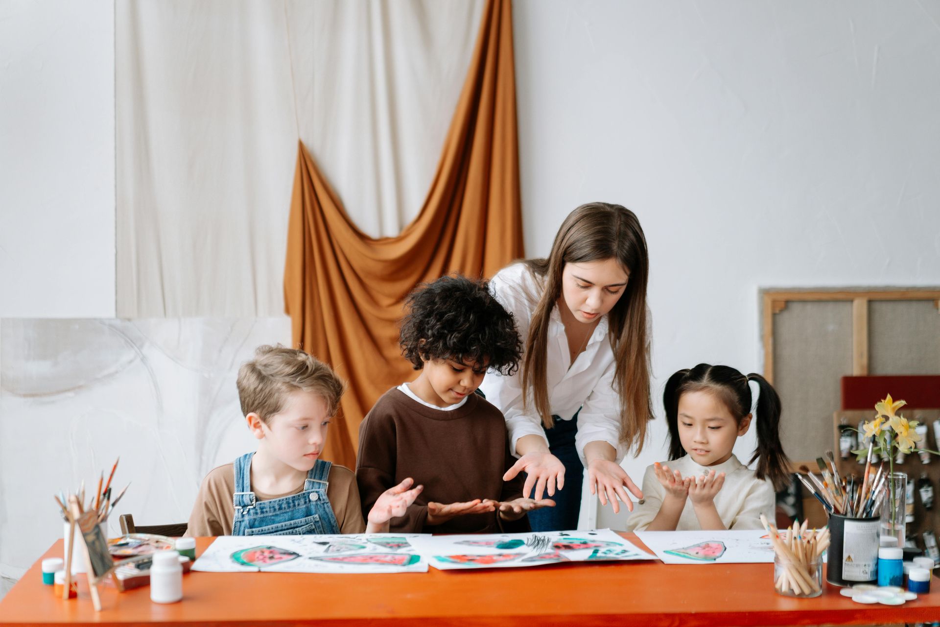 Teacher helps four children with art project at a red table; brown cloth backdrop.