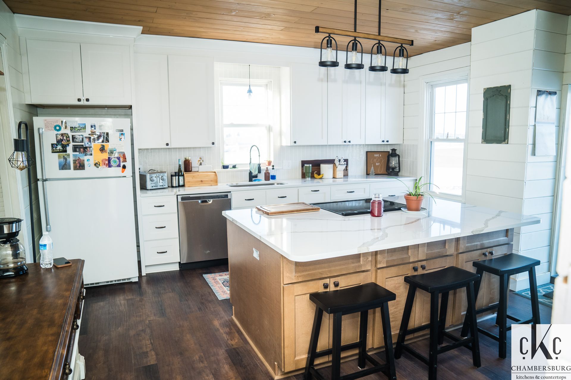 A kitchen with white cabinets, stools, a refrigerator and a large island.
