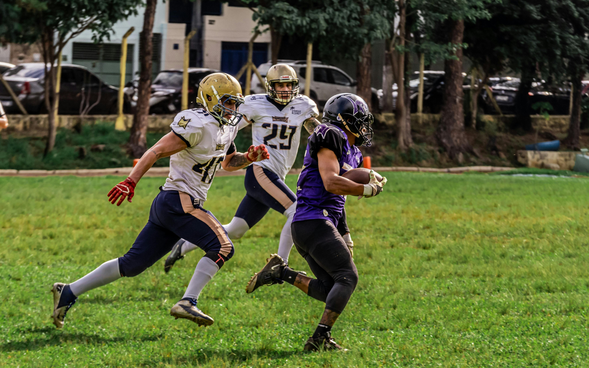 Football players in action on a grassy field; one runs with the ball, pursued by two opponents.