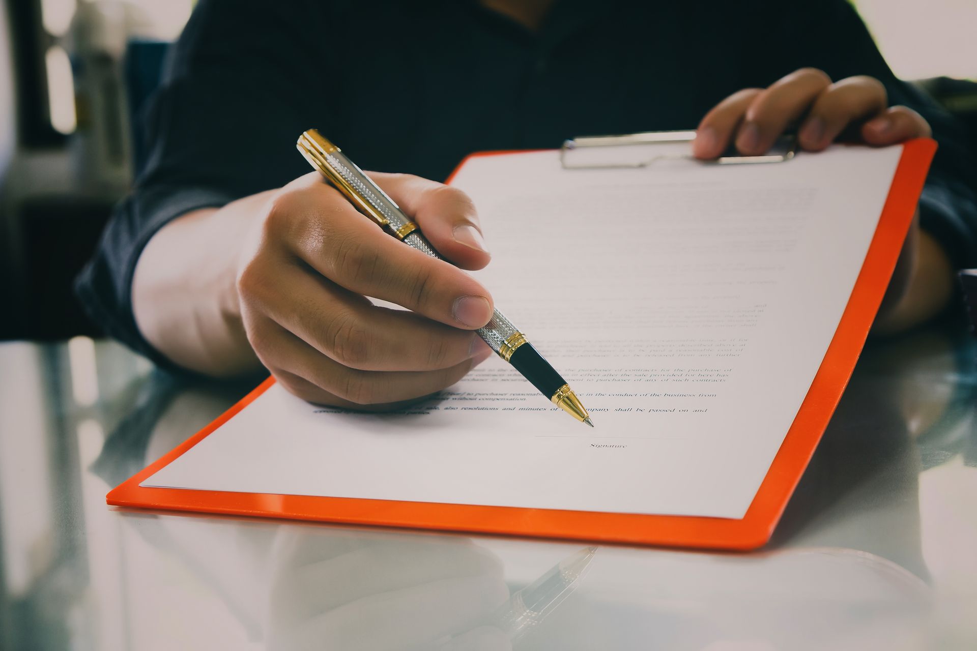 Hand holding a pen over a clipboard with a blank form and orange border.