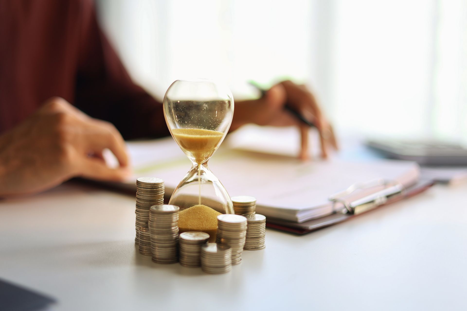 Hourglass and coin stacks on a desk beside a blurred person signing documents.
