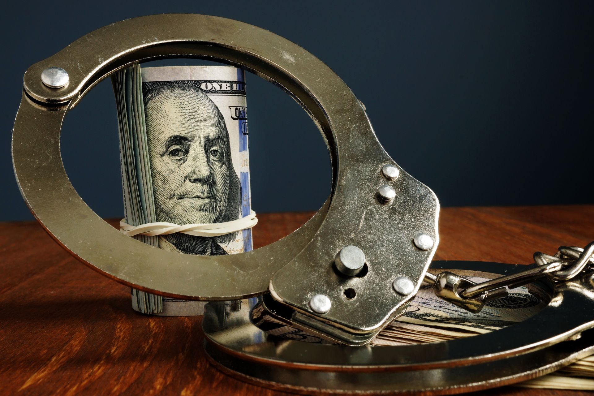Handcuffs around a rolled $100 bill on a wooden surface, symbolizing financial crime or arrest.