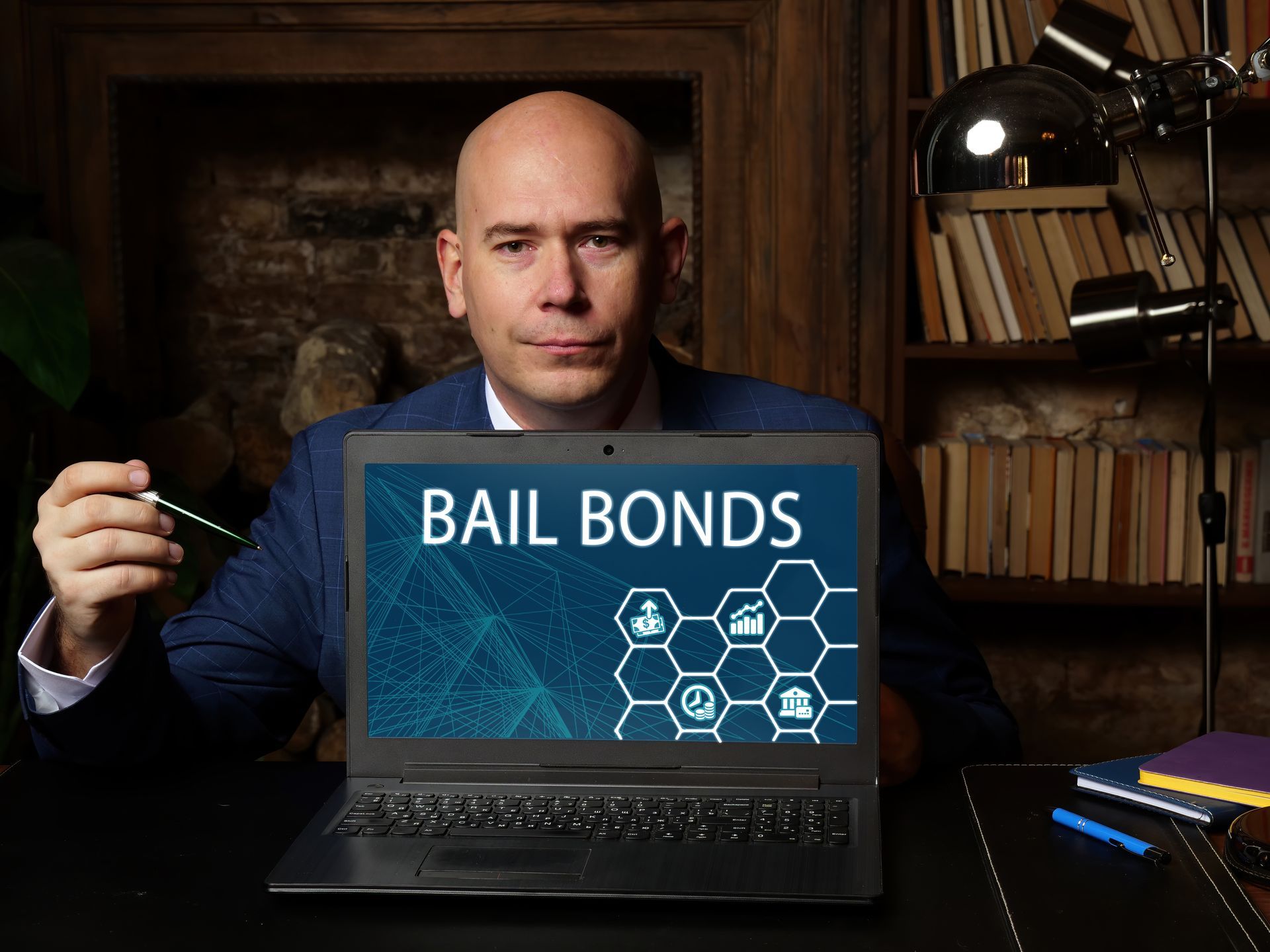 Man seated at desk holding a pen, laptop screen reading “BAIL BONDS” in a dim office setting.