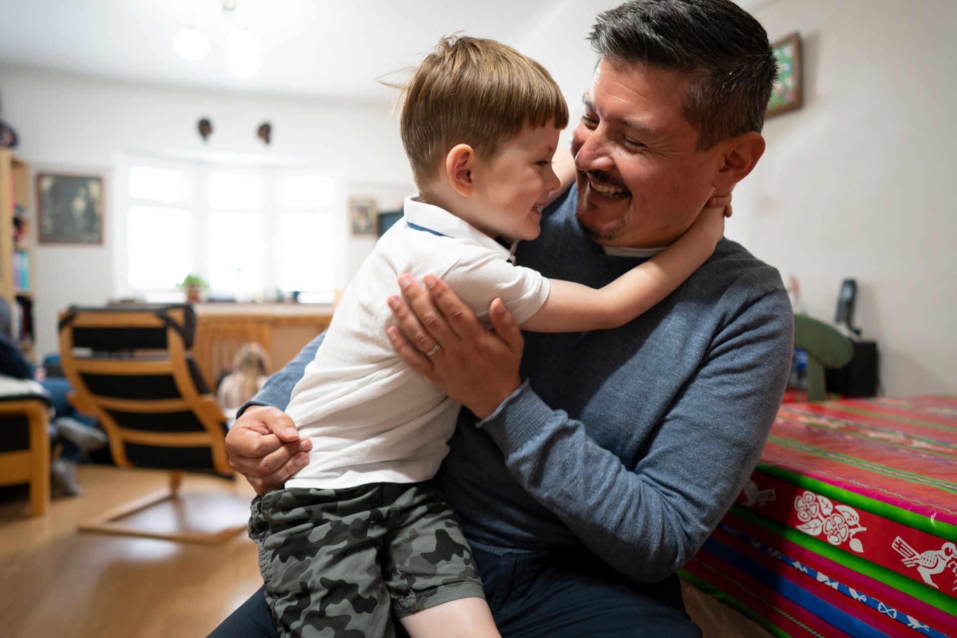 Boy hugging smiling man indoors beside a red patterned gift box.