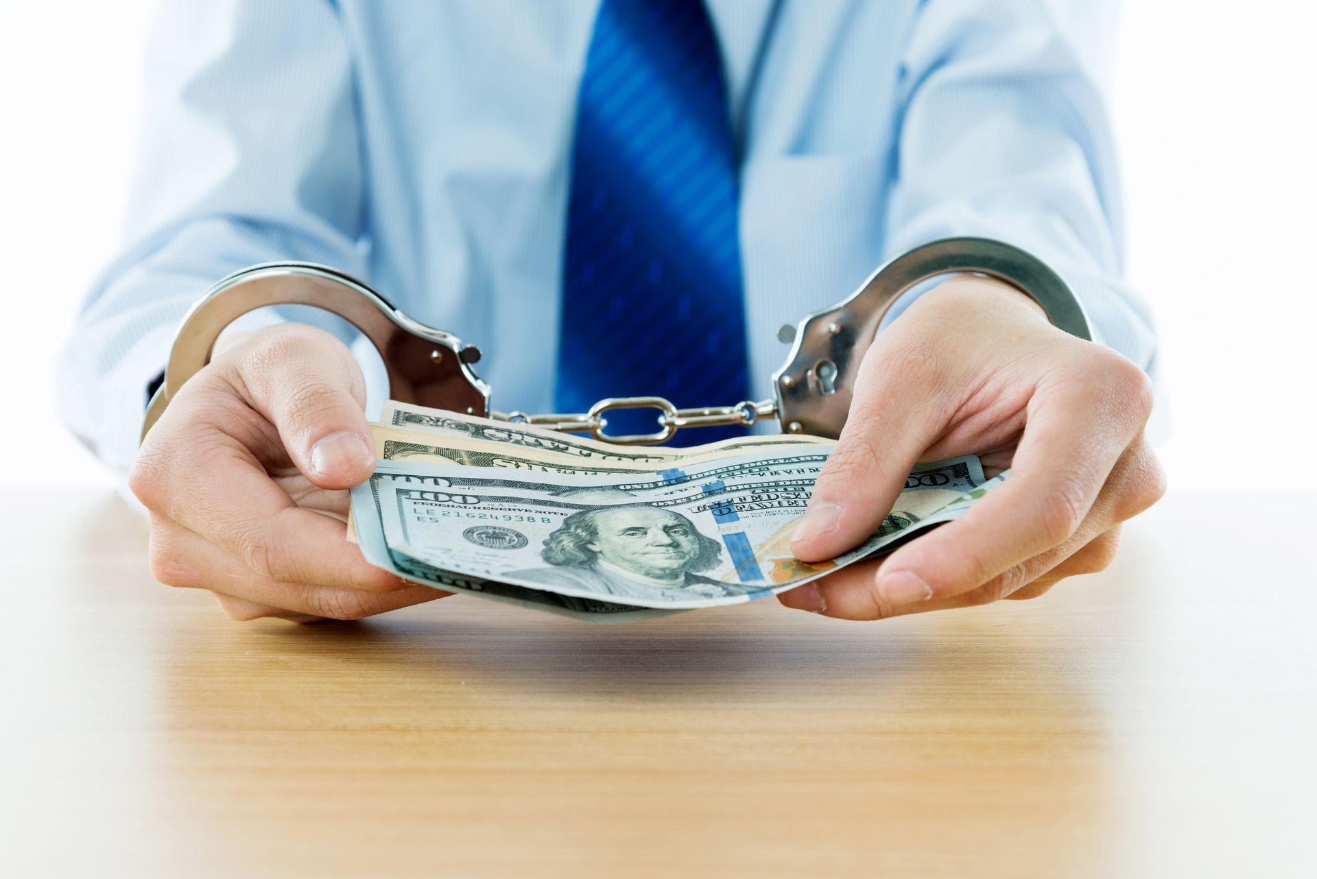 Handcuffed person in a white shirt and blue tie holding U.S. dollar bills at a table.