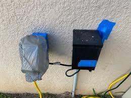 Two black electrical boxes, one with a blue tape and the other covered in gray plastic, mounted on an off-white wall.