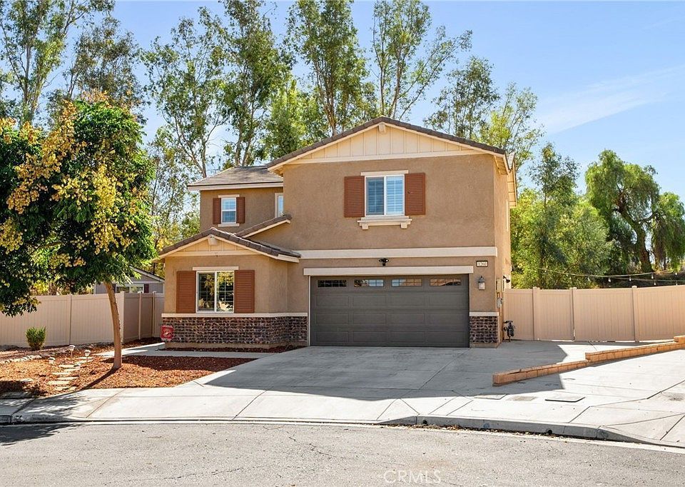 Two-story beige suburban house with a brown garage, a stone base, and a tan fence under a bright blue sky.