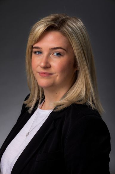 A woman with blonde hair and blue eyes is wearing a black jacket and a white shirt.