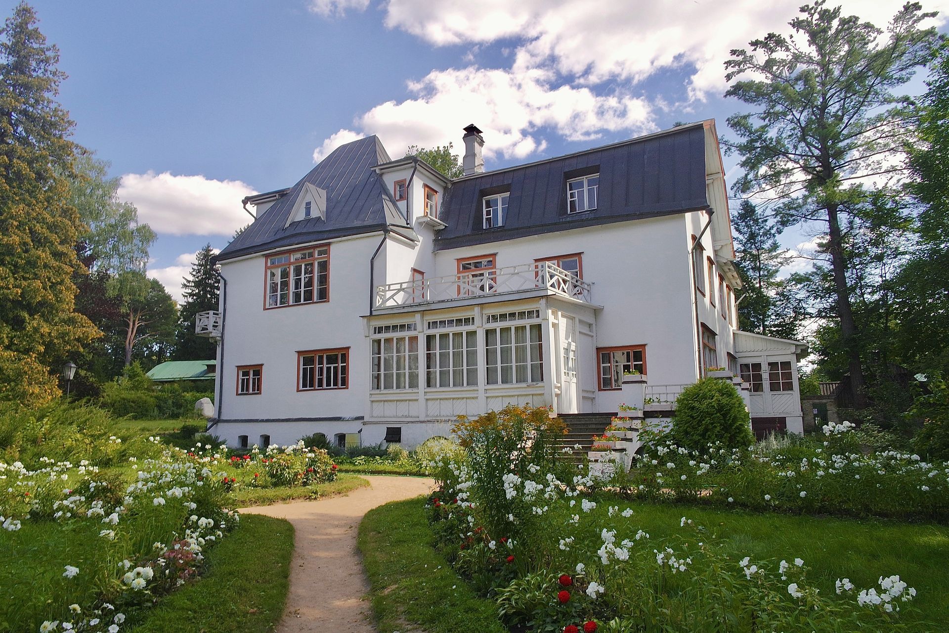 A large white house with a black roof is surrounded by trees and flowers