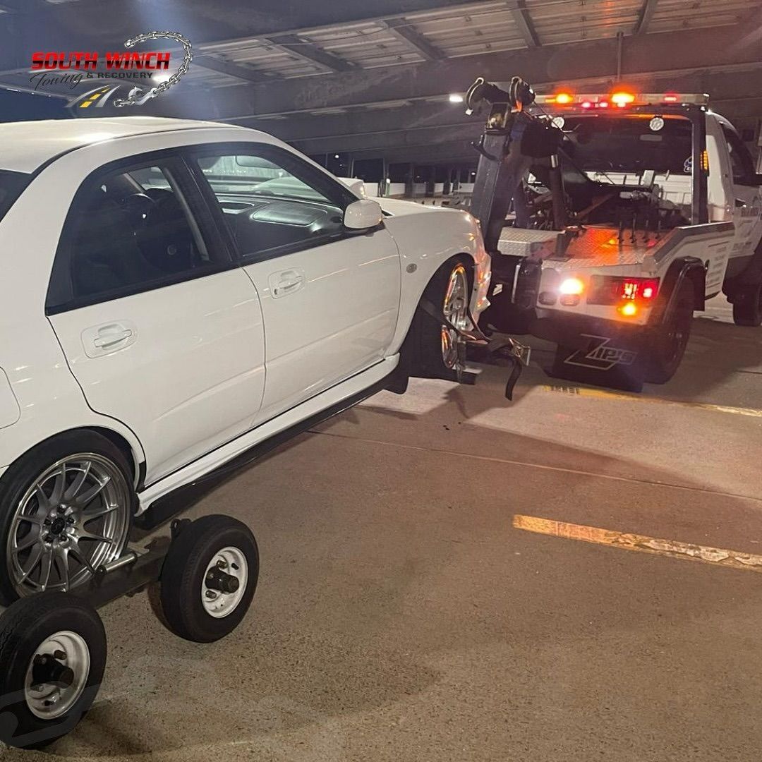 A white car is being towed by a tow truck in a parking lot.