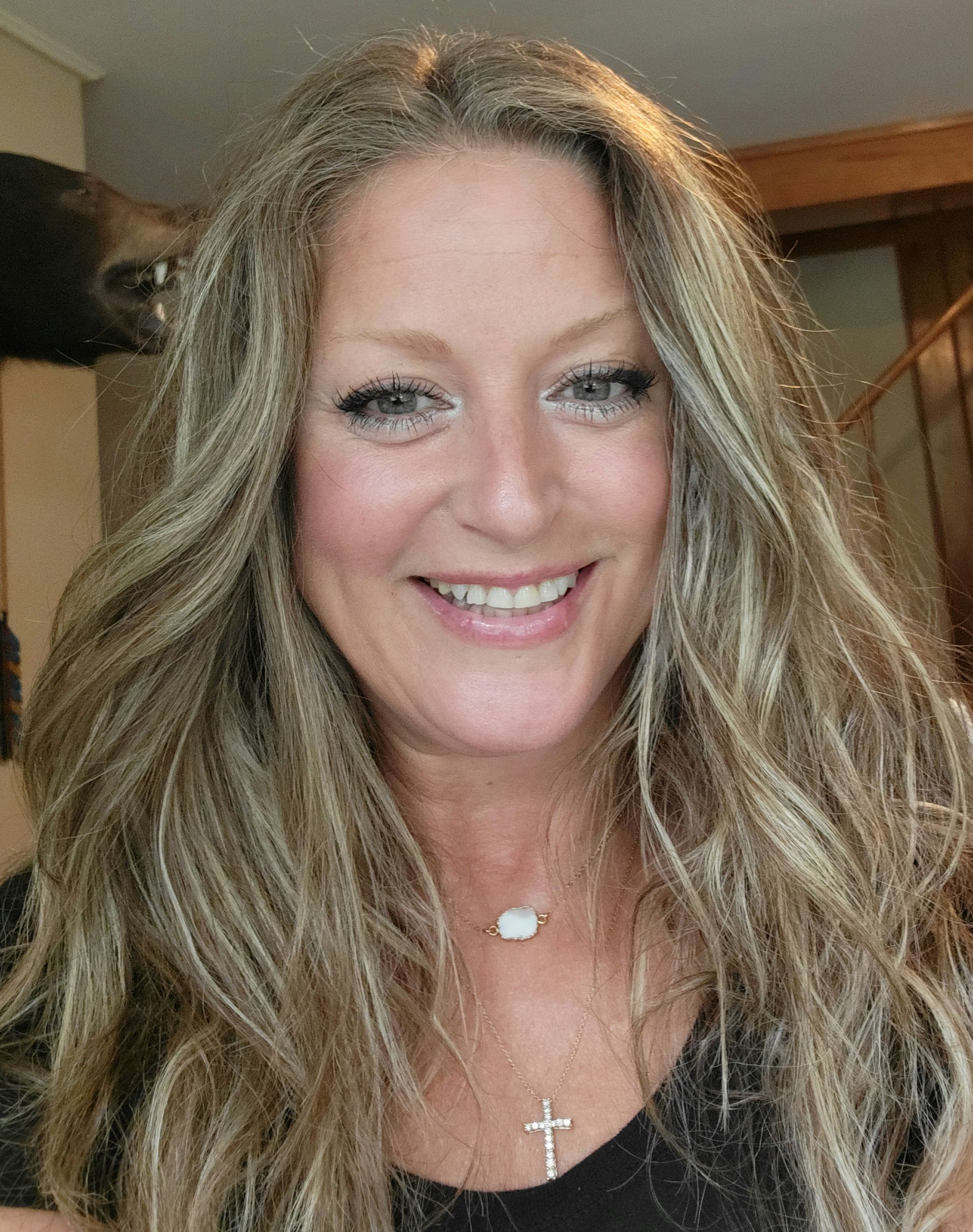 A woman with long blonde hair is smiling and wearing a cross necklace.