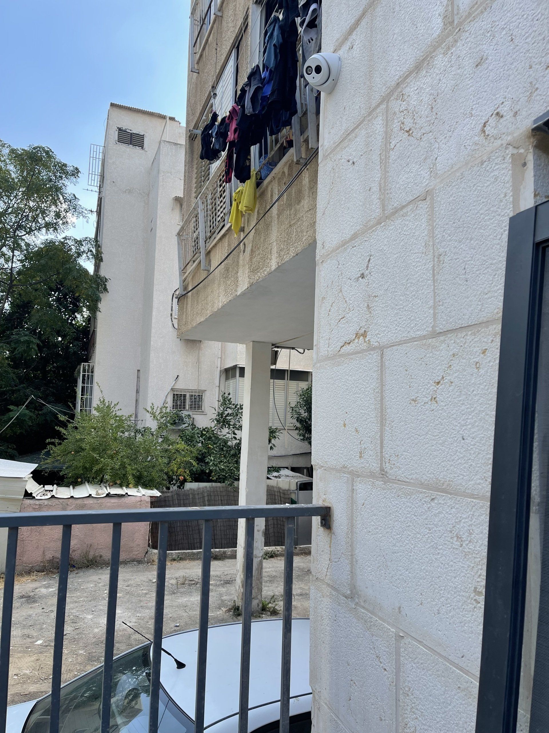 מכונית לבנה חונה מול בניין עם בגדים תלויים במרפסת.