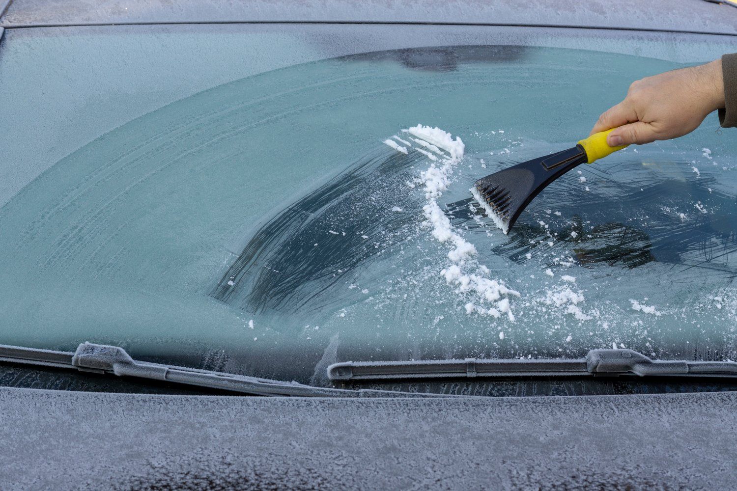 A person is cleaning the windshield of a car with an ice scraper.