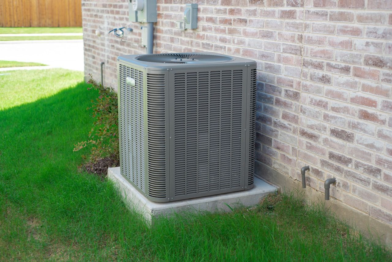 A grey residential air conditioning unit sitting on a concrete pad next to a brick house exterior.