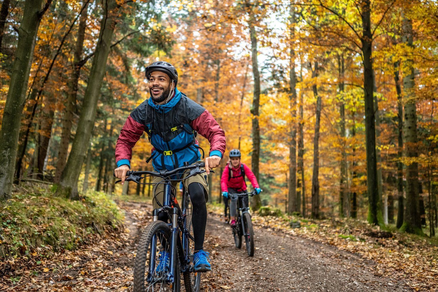 A man and a woman are riding bicycles down a dirt road in the woods.