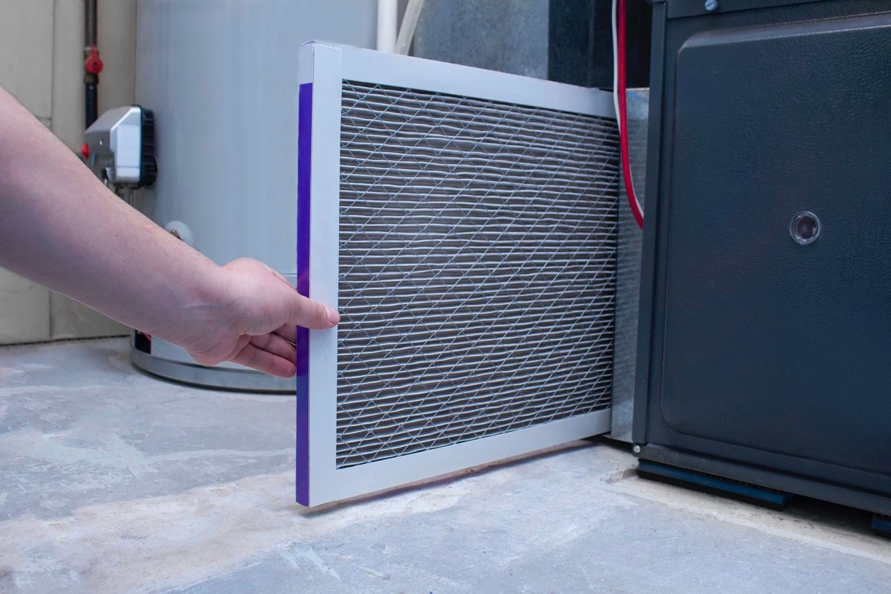 A hand sliding a rectangular HVAC air filter into a slot on a furnace unit next to a water heater.