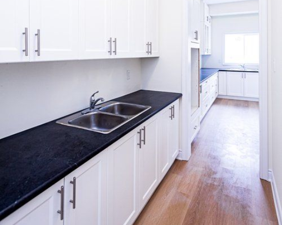 White butler's kitchen with dark countertops, a double sink, and cabinets. Hardwood floors and a window are visible in the background.