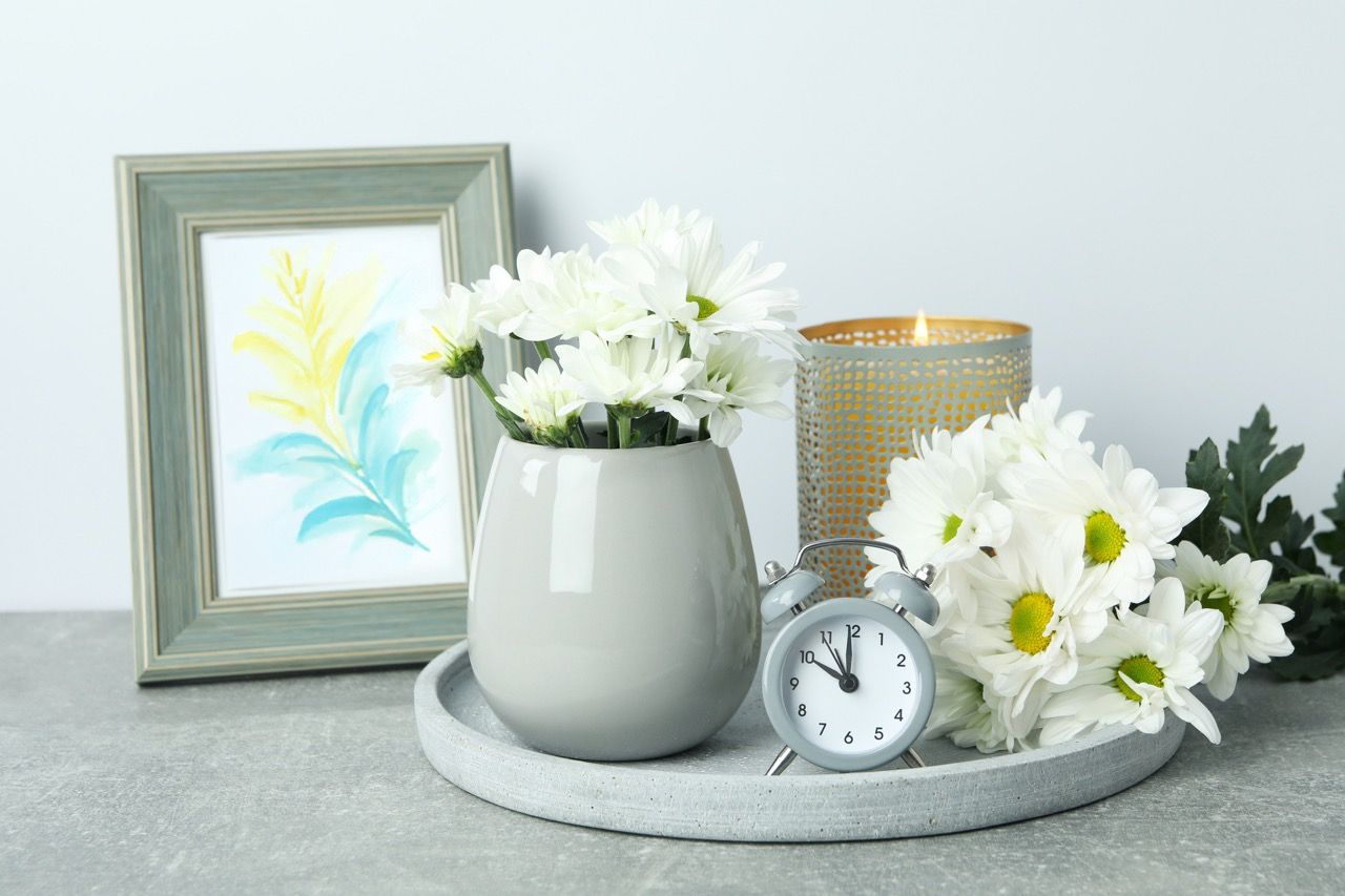 A grey tray holding a grey vase of white chrysanthemums, a small alarm clock, and a lit candle beside a framed watercolor.